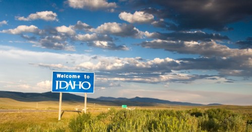A blue sign reading, 'Welcome to Idaho,' sits on a yellow grass field with mountains far in the background and dramatic grey clouds overhead.