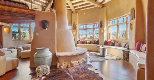 Sun-filled adobe living room in Arroyo Seco, New Mexico, with curved seating, exposed beams, and a sculptural fireplace inspired by the communal living spaces of Morocco.