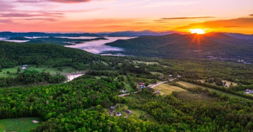 The sun sets on a wide expanse of green valleys covered in forests and small towns with clouds clung low to the hills.