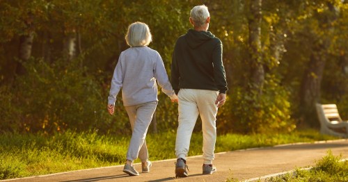 Married retired couple walking together on a sunny summer evening. From behind a retired man and woman in sports clothes holding hands walking on a park walkway among green trees in Minnesota.