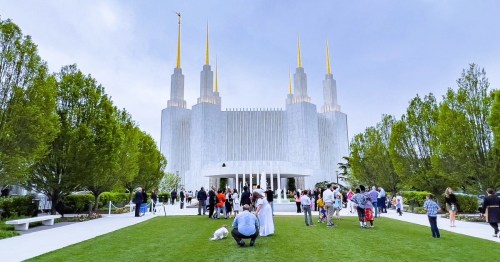 View of the Washington DC Temple, with six spires topped with golden finials, with a crowd of people on the manicured lawn.