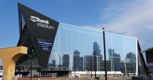 The striking, angular glass exterior of U.S. Bank Stadium in Minneapolis reflects the surrounding city buildings and blue sky, highlighting its modern design as the home of the Minnesota Vikings.