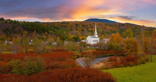 A small town sits shrouded by a forest turning orange and red with fall colors but one white church stands out prominently with the sunset striking the mountain behind.