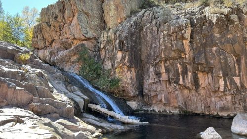 A serene waterfall cascades over rocky cliffs into a calm pool, surrounded by trees and clear blue skies.