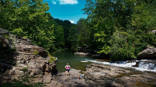 A serene river scene with lush green trees, rocky banks, and two children exploring near the water under a blue sky.