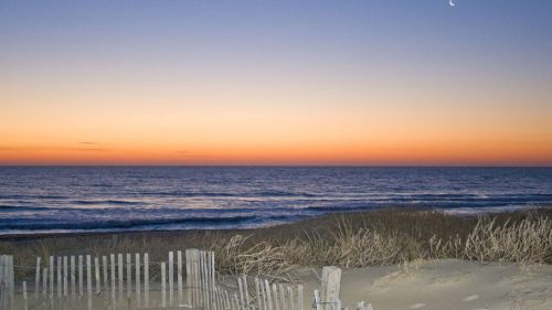 A serene beach at sunset, with gentle waves, a colorful sky, and a crescent moon above the horizon.