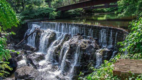 A serene waterfall cascades over rocks, surrounded by lush greenery, with a bridge in the background.