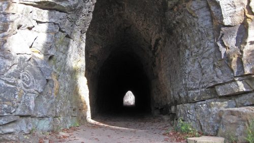A dark stone tunnel with a lighted exit, surrounded by rugged rock walls and scattered leaves on the ground.