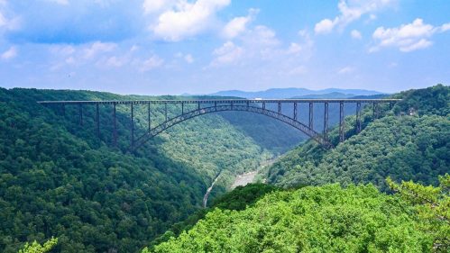 A tall bridge spans a lush green valley, surrounded by mountains under a blue sky with fluffy clouds.