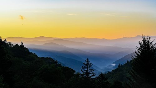 A serene mountain landscape at sunset, with layered hills and a gradient sky in shades of orange and blue.