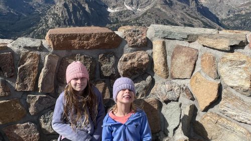 Two girls in winter hats kneel by a stone wall, smiling with mountains in the background on a sunny day.