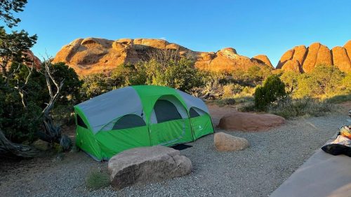 A green and gray tent set up on rocky ground, surrounded by shrubs and mountains under a clear blue sky.