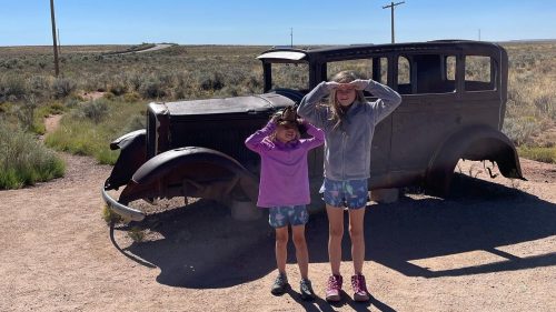 Two girls stand beside an old, rusty car in a desert landscape under a clear blue sky.