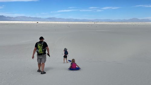 A man and two children walk on a vast white sand landscape under a blue sky with clouds.