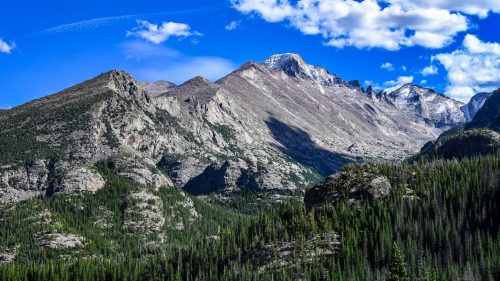 A scenic view of rugged mountains under a bright blue sky with fluffy clouds, surrounded by lush green forests.