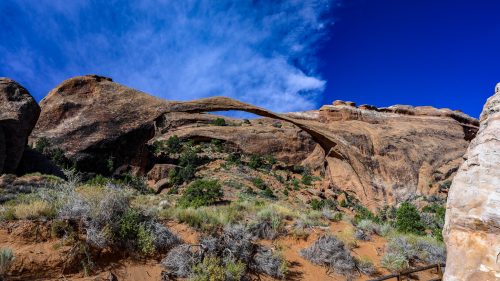 A natural stone arch rises above a desert landscape under a bright blue sky with scattered clouds.