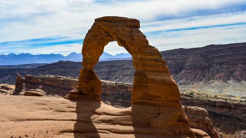A natural stone arch stands against a blue sky, surrounded by rugged mountains and desert landscape.
