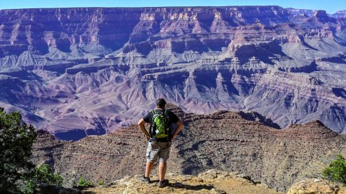 A hiker stands on a rocky ledge, gazing at the vast, colorful landscape of the Grand Canyon under a clear blue sky.