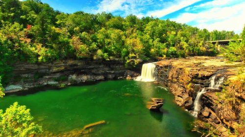 A scenic view of a waterfall cascading into a green pool, surrounded by lush trees and a blue sky with clouds.