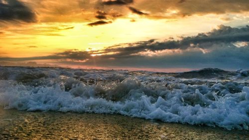Waves crashing on a beach at sunset, with vibrant orange and yellow hues in the sky and dramatic clouds.