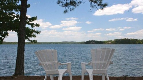 Two white Adirondack chairs facing a calm lake under a blue sky with fluffy clouds, surrounded by trees.
