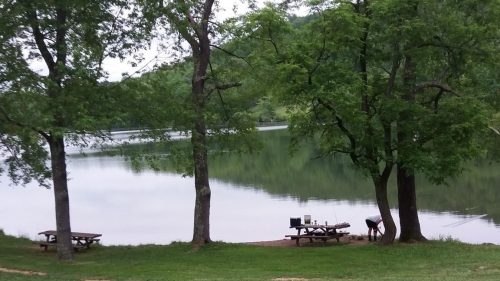 A serene lakeside scene with picnic tables under trees, reflecting calm water and greenery in the background.