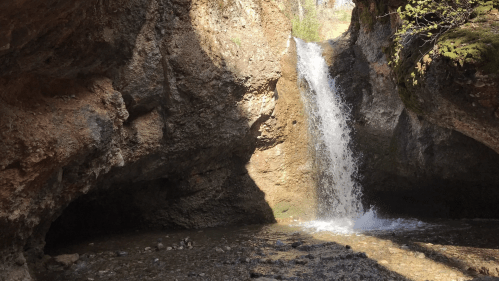 A serene waterfall cascades between rocky cliffs, surrounded by greenery and sunlight filtering through the trees.