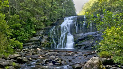 A serene waterfall cascades over rocks, surrounded by lush green trees and a tranquil stream below.
