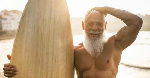 A fit senior citizen with a white beard, holding a vintage surfboard.