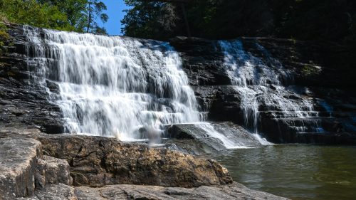 A serene waterfall cascading over rocky ledges into a calm pool, surrounded by lush greenery and trees.