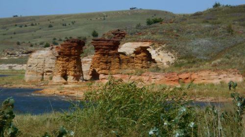 Rock formations rise above a river, surrounded by grassy hills and a clear blue sky.