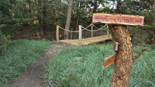 A wooden sign points to "Chetolah" near a grassy path leading to a small bridge in a wooded area.