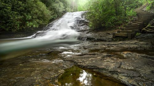 A serene waterfall cascades over rocks, surrounded by lush greenery and a misty atmosphere, with stone steps nearby.