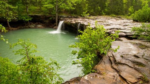 A serene waterfall cascades into a turquoise pool, surrounded by lush greenery and rocky terrain.