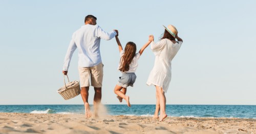 A family on the beach, seen from behind. A mother and father are standing facing the beach as they playfully lift their young daughter up by her arms.