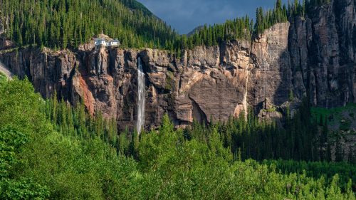 A waterfall cascades down a rocky cliff surrounded by lush green trees and mountains under a cloudy sky.