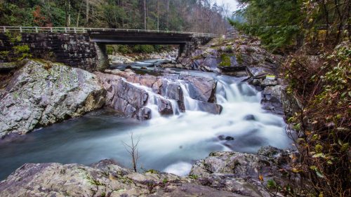 A serene river flows over rocks beneath a bridge, surrounded by lush greenery and misty mountains in the background.
