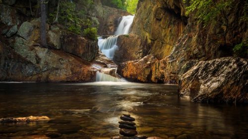 A serene waterfall cascades over rocks into a calm pool, with a stack of stones balanced on a nearby boulder.