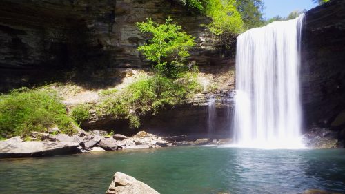 A serene waterfall cascades into a clear pool, surrounded by lush greenery and rocky terrain.