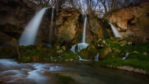 A serene waterfall cascades over rocky cliffs, surrounded by lush greenery and a tranquil stream.