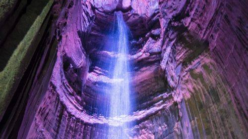 A vibrant waterfall cascades down a rocky, illuminated cavern, showcasing colorful lighting and textured walls.