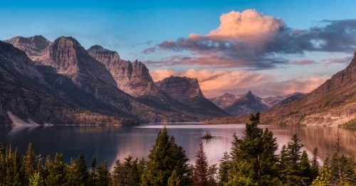A calm lake with rugged mountains and tall pine trees surrounding it at sunset.