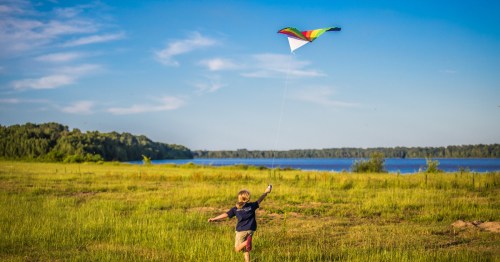 A boy runs in a green field with a kite flying overhead with water in the distant background.
