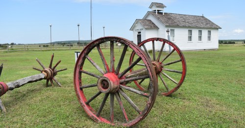 A red wagon wheel stands in green grass in front of a white schoolhouse.