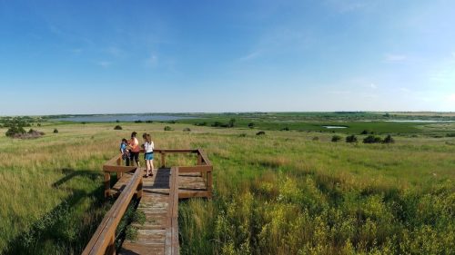 Three people stand on a wooden boardwalk overlooking a vast green landscape and a serene body of water under a clear sky.