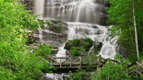 A serene waterfall cascades down rocky cliffs, surrounded by lush green trees and a wooden bridge in the foreground.