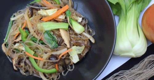 A close-up of a black plate of noodles with stir-fried vegetables. Cabbage, dry noodles, and an onion are visible on the side of the plate.