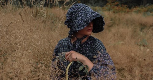A girl in a Prairie-style blue bonnet and dress stands in a field.