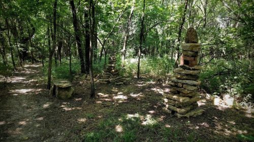 Two stone cairns stand among trees in a lush, green forest, with a dirt path visible on the left.