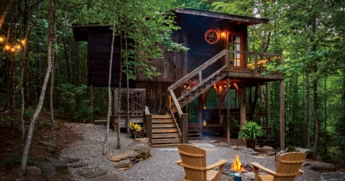 A rustic treehouse surrounded by lush green trees and foliage. Wooden chairs sit near the base of the stairs, next to a roaring fire.